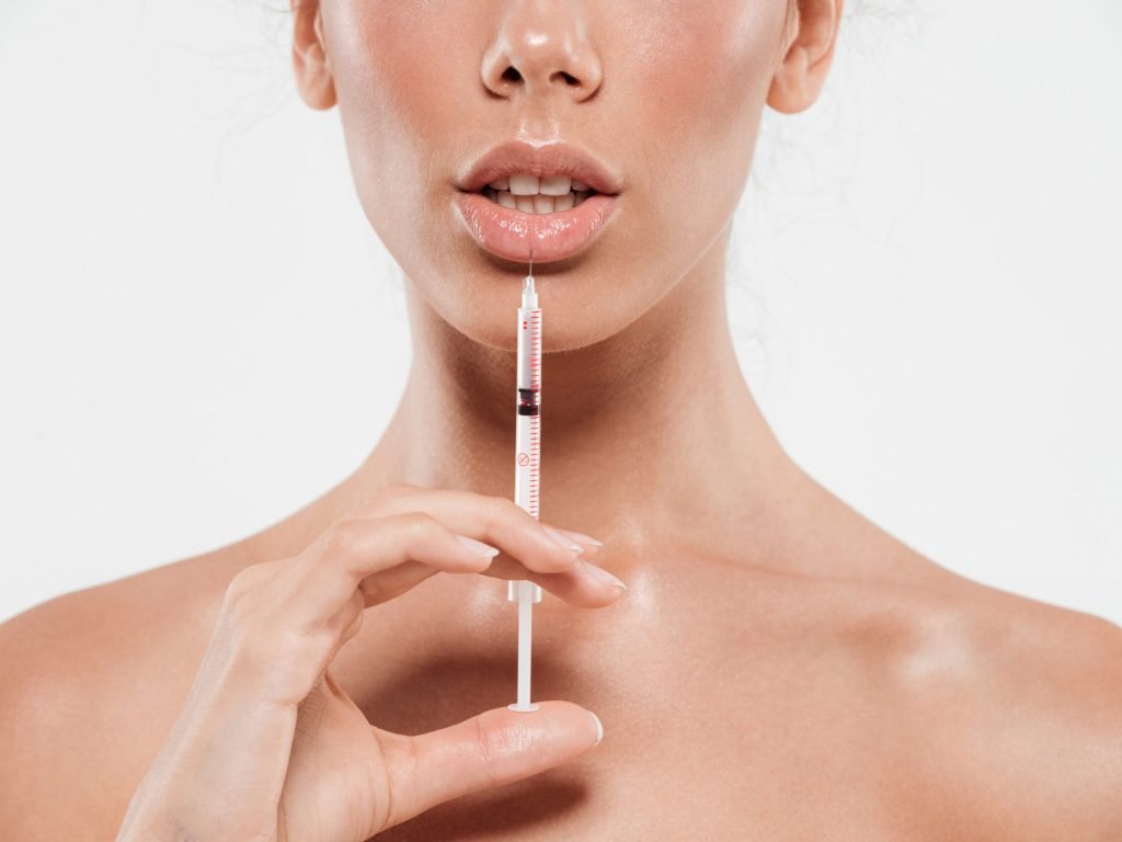 Cropped image of young female face and a hand holding a syringe and making injection in her lip isolated over white background