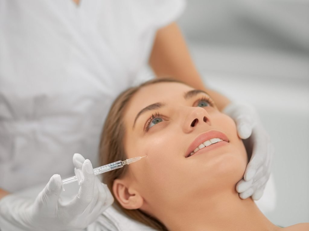 Close up of smiling young brunette woman on procedure injection anti-aging skin in beautician. Concept of process injection for improve the condition face.